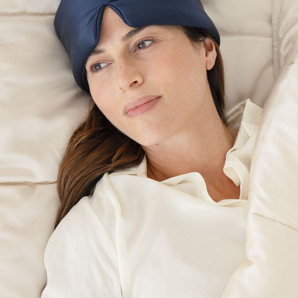 A woman in a white shirt, long brown hair flowing, lies in bed partially covered by the drowsysleepco Dusty Gold Silk Weighted Blanket, with a navy sleep mask pushed up on her forehead, gazing thoughtfully.