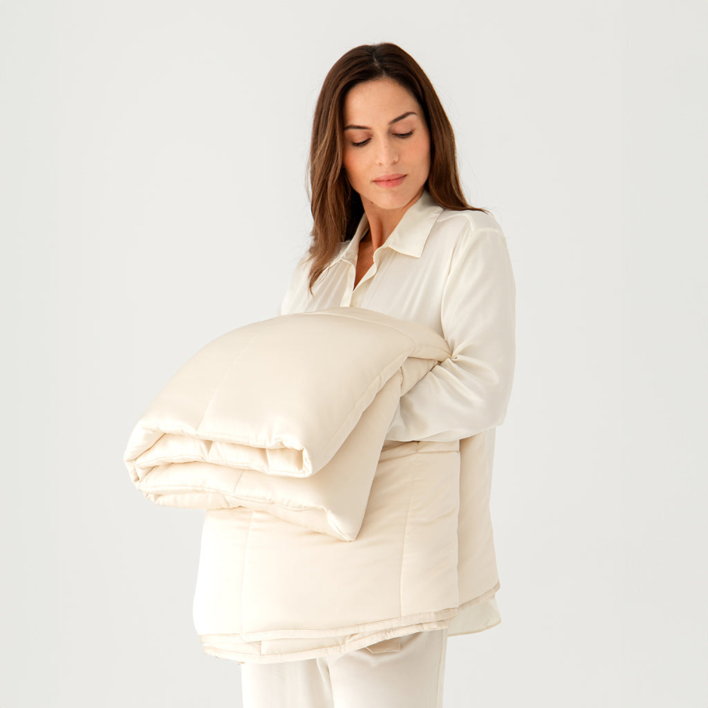 A woman in light pajamas holds a folded Dusty Gold Silk Weighted Blanket by drowsysleepco against her chest, standing before a plain white background.