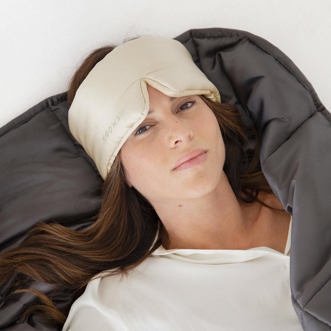 A woman with long brown hair relaxes on her back, wearing a cream sleep mask and white shirt, covered by the drowsysleepco Moonlight Shadow Silk Weighted Blanket in dark gray.