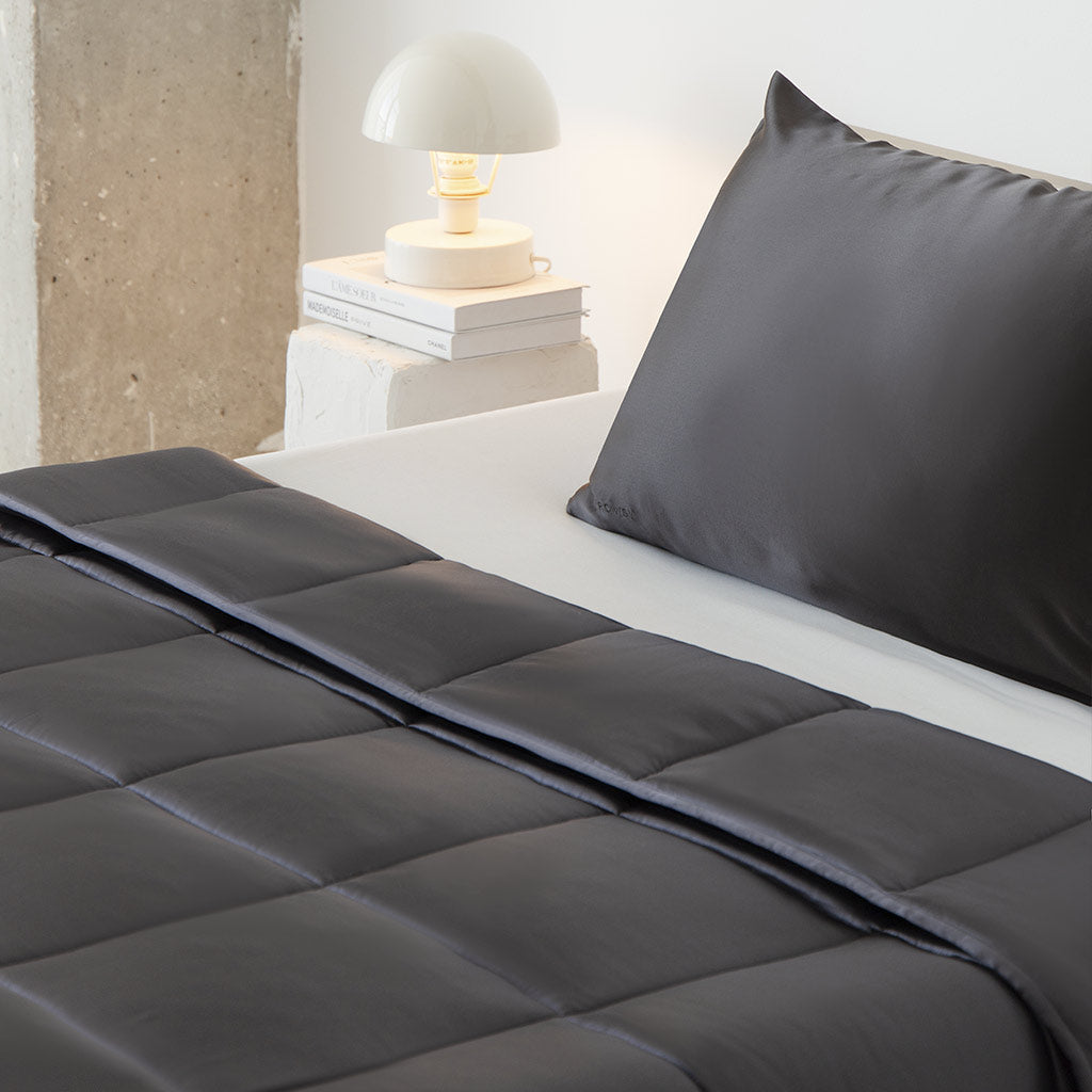 A neatly made bed with dark gray bedding and the drowsysleepco Moonlight Shadow Silk Weighted Blanket, beside a white nightstand topped with stacked books and a round white lamp, set against a concrete pillar and white wall.