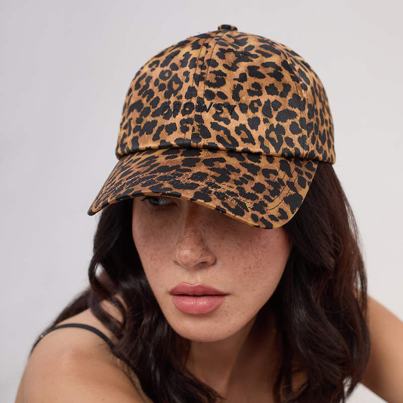 A woman with dark hair wears the Drowsy Silk Cap - Hotel Paradiso Wild Thing by Drowsy, a brown and black leopard print cap for hair protection, along with a black strap top. She looks slightly downward with a neutral expression.