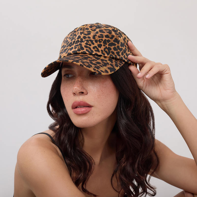 A woman with long dark hair and freckles poses against a light background, wearing the Drowsy Silk Cap - Hotel Paradiso Wild Thing by Drowsy for hair protection, paired with a black strap top and touching the brim of her cap.