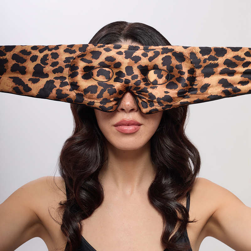 A woman with long, wavy dark hair holds the us.drowsysleepco Hotel Paradiso Wild Thing Eyelash Protecting Mask—a wide leopard-print sleep mask with eye holes—over her face against a plain background.