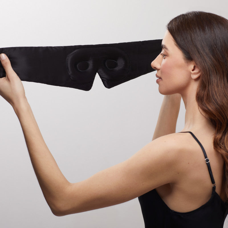 A woman with long brown hair, wearing a black camisole, holds up the us.drowsysleepco Black Jade - Eyelash Protecting Mask with eye indentations, preparing to put it on against a neutral background.