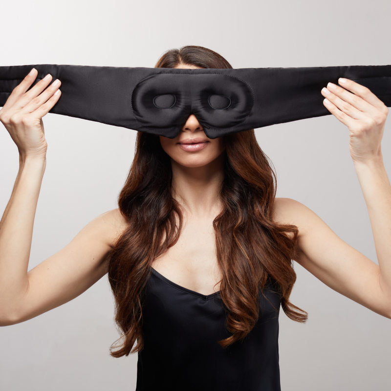 A woman with long brown hair in a black camisole holds up the us.drowsysleepco Black Jade - Eyelash Protecting Mask, a large silk blackout mask with eye holes, preparing to put it on against a plain light background.