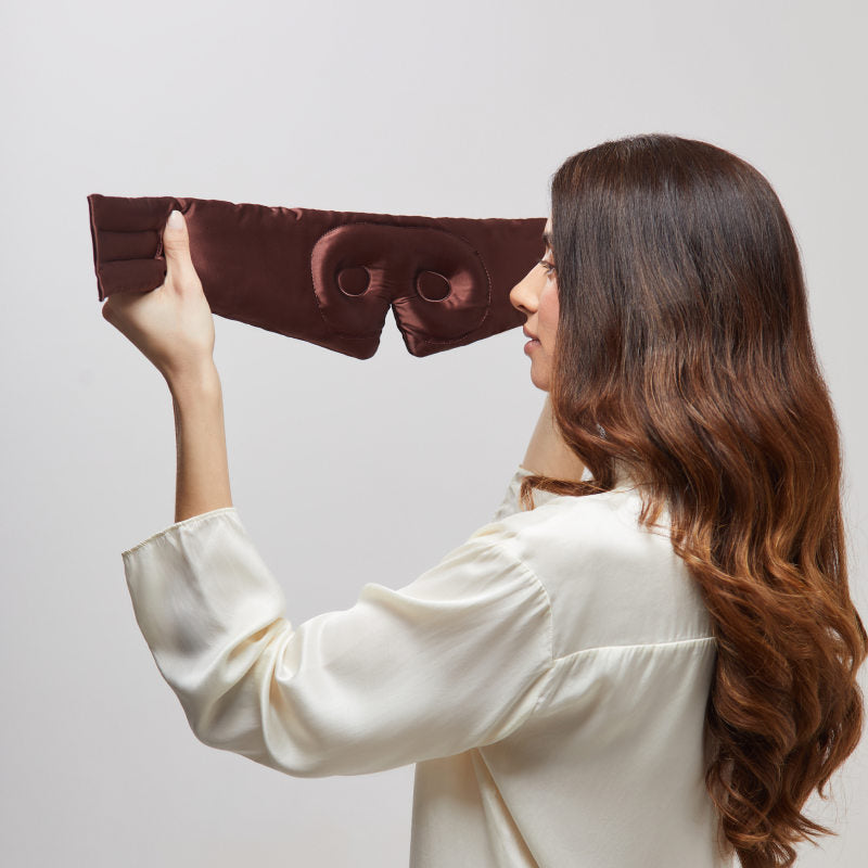 A woman with long brown hair in a white blouse holds up the us.drowsysleepco Cocoa Glow - Eyelash Protecting Mask, admiring its eye indentations and lash-saving design, set against a plain light background.
