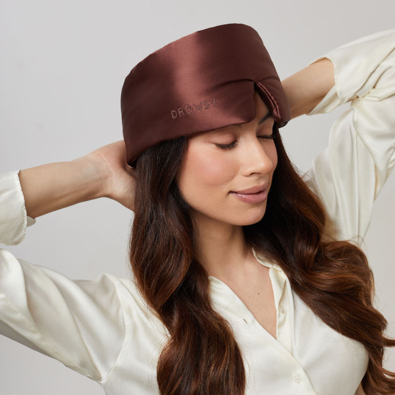 A woman with long brown hair wears the us.drowsysleepco Cocoa Glow Eyelash Protecting Mask and a cream blouse, gently touching the back of her head and smiling with her eyes closed.