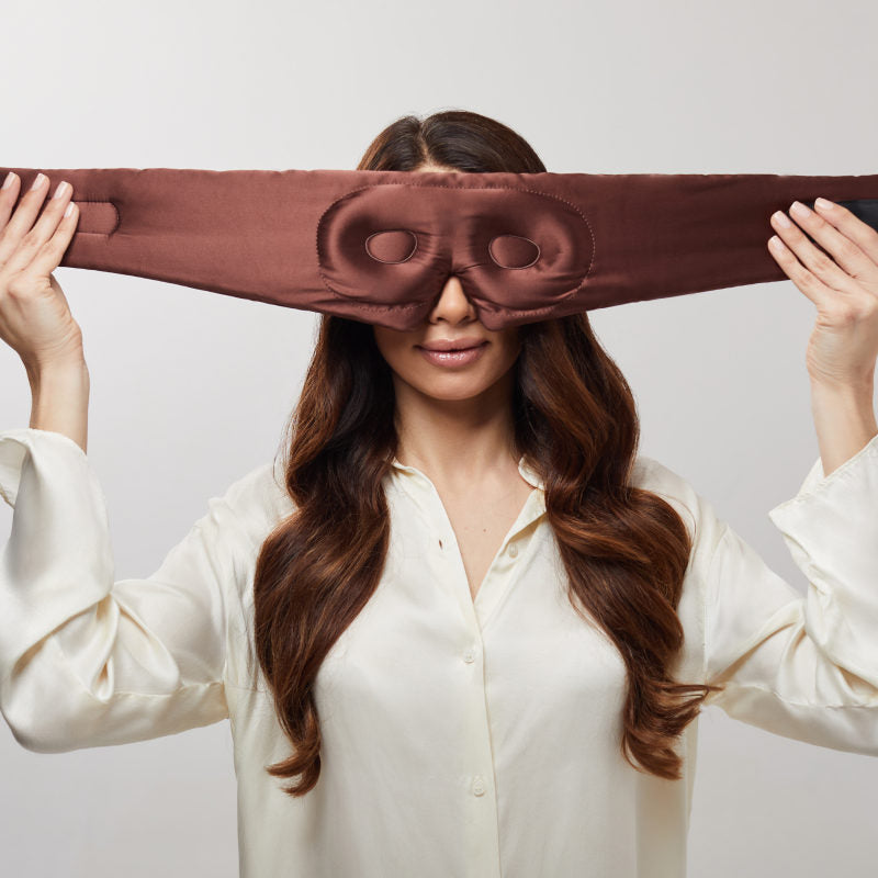 A woman with long brown hair and a white blouse holds the us.drowsysleepco Cocoa Glow - Eyelash Protecting Mask over her eyes, preparing to put it on for comfortable, eyelash-safe rest.