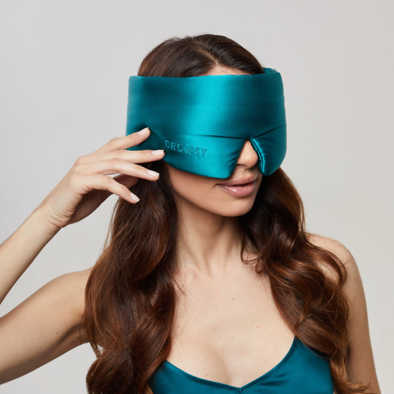 A woman with long brown hair wears the us.drowsysleepco Green Sapphire - Eyelash Protecting Mask with contoured eyecups, matching her teal top, and holds one side of the mask against a light background.