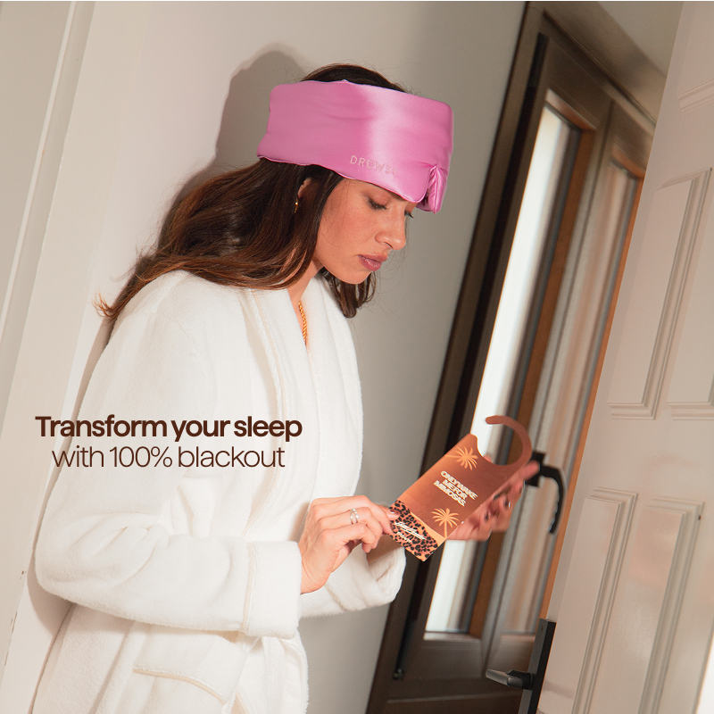 A woman in a white robe wears the pink Hotel Paradiso Heatwave Sleep Mask by us.drowsysleepco on her forehead, standing by a door holding a brown box. Text reads: Transform your sleep with 100% blackout.