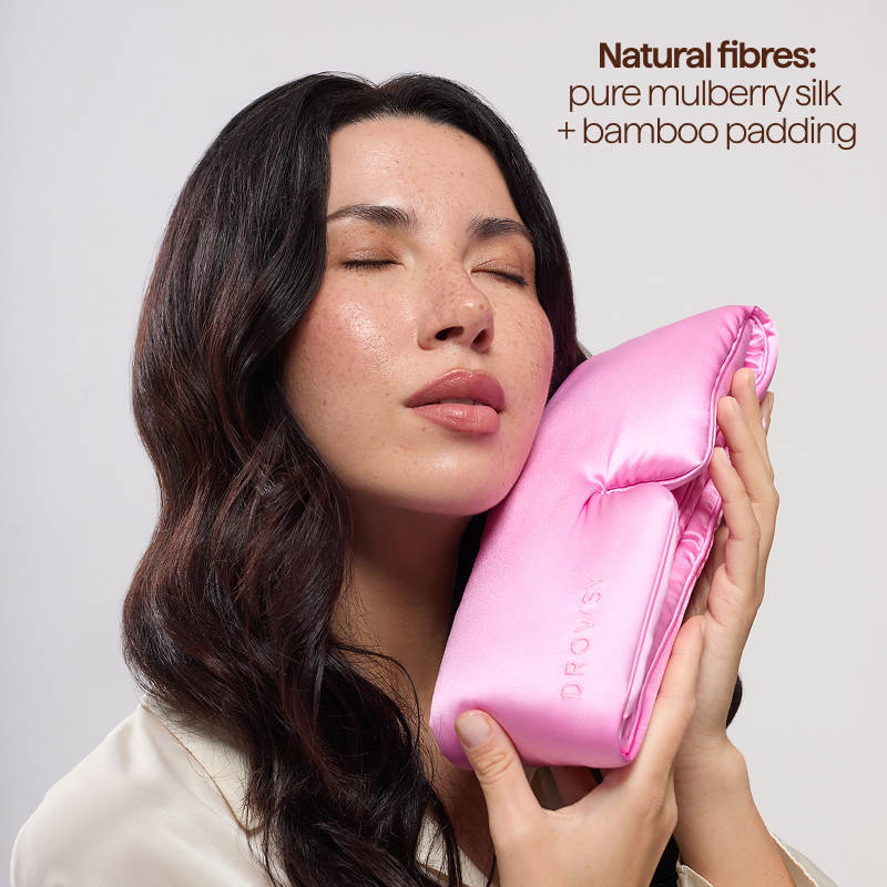 A woman with closed eyes holds a pink silk pillow against her cheek. Text: Hotel Paradiso Heatwave Sleep Mask by us.drowsysleepco—pure mulberry silk and bamboo padding for the comfort of a luxury sleep mask and blissful rest.
