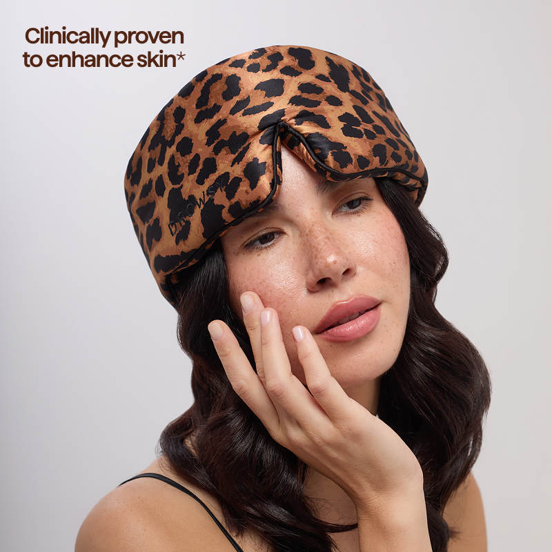 A woman with long wavy hair wears the Hotel Paradiso Wild Thing Sleep Mask by us.drowsysleepco, featuring a leopard print. Text in the top left corner reads, "Clinically proven to enhance skin hydration." The background is plain light beige.