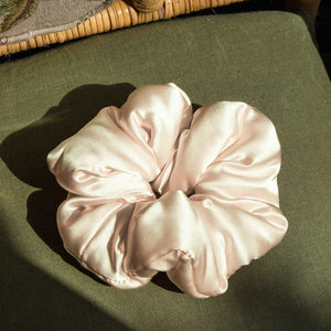 The Champagne Gold Padded Scrunchie by us.drowsysleepco sits on an olive green upholstered surface, sunlight casting soft shadows—an oversized, luxe accessory perfect for adding hair volume and enhancing your sleep routine.