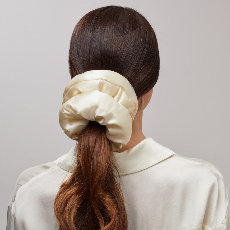A woman with long brown hair in a low ponytail wears the us.drowsysleepco Champagne Gold Padded Scrunchie and a cream satin blouse, photographed from behind. This oversized scrunchie boosts hair volume and serves as a chic sleep accessory.