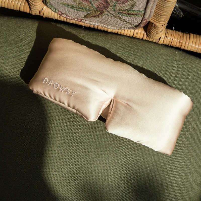 A Champagne Gold silk eye mask by us.drowsysleepco rests on green fabric, ideal for beauty sleep, with part of a woven basket visible in the background.