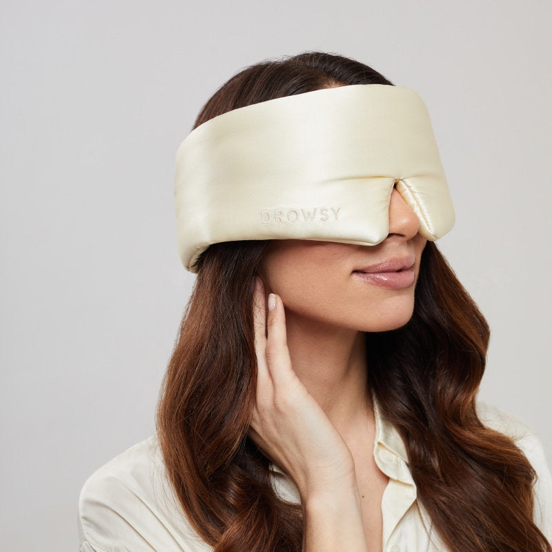 A woman with long brown hair wears the Champagne Gold silk eye mask by us.drowsysleepco, embroidered with DROWSY. She smiles gently while touching her face, enjoying beauty sleep and optimal skin hydration.