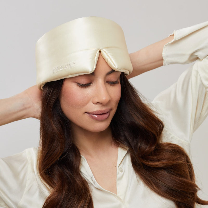 A woman with long, wavy brown hair wears the us.drowsysleepco Champagne Gold silk eye mask on her forehead and a matching satin shirt, relaxing with closed eyes and enjoying restful beauty sleep.