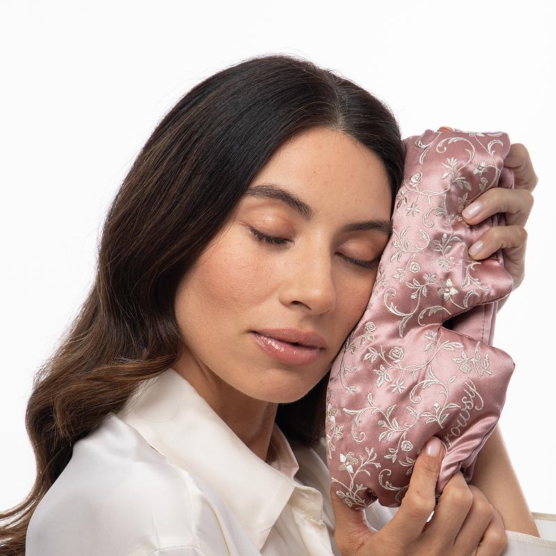 A woman with her eyes closed rests her cheek against a pink Pride & Prejudice - English Rose Eyelash Protecting Sleep Mask embroidered with floral vines and the word Drowsy.