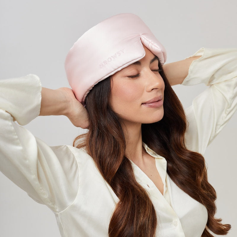 A woman with long brown hair wears the us.drowsysleepco Petal Pink silky blackout eye mask and a white blouse. She closes her eyes, smiling gently in a relaxed pose with her hands raised behind her head.