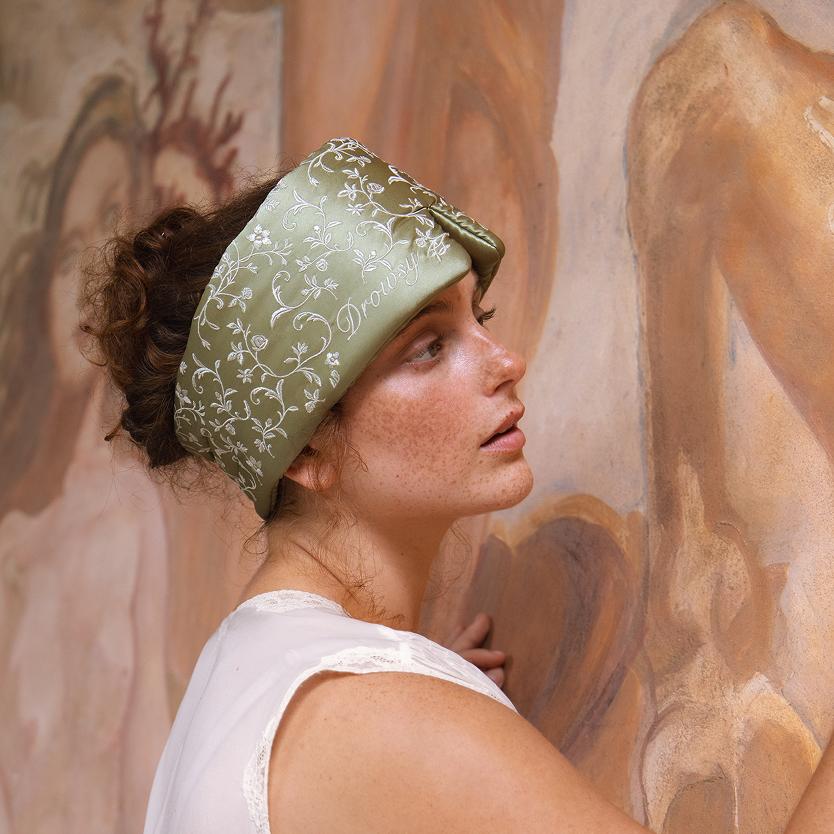 A person wearing a sage green Pride & Prejudice - Sage Sleep Mask featuring white floral patterns and embroidery.