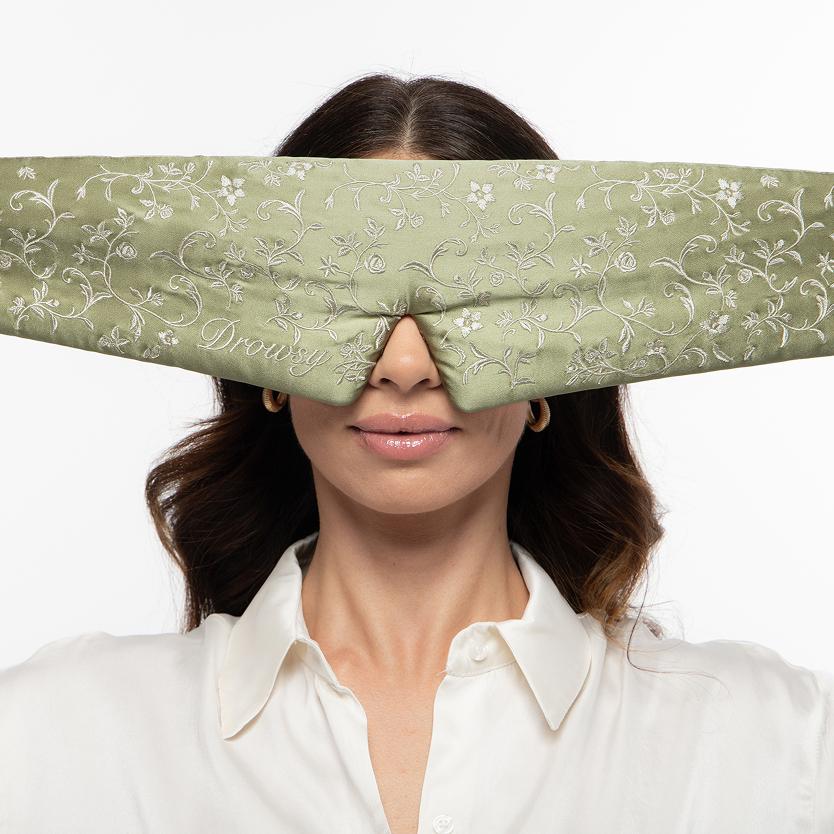 A person wearing a sage green Pride & Prejudice - Complete collection original mulberry silk sleep mask embroidered with a white floral pattern.