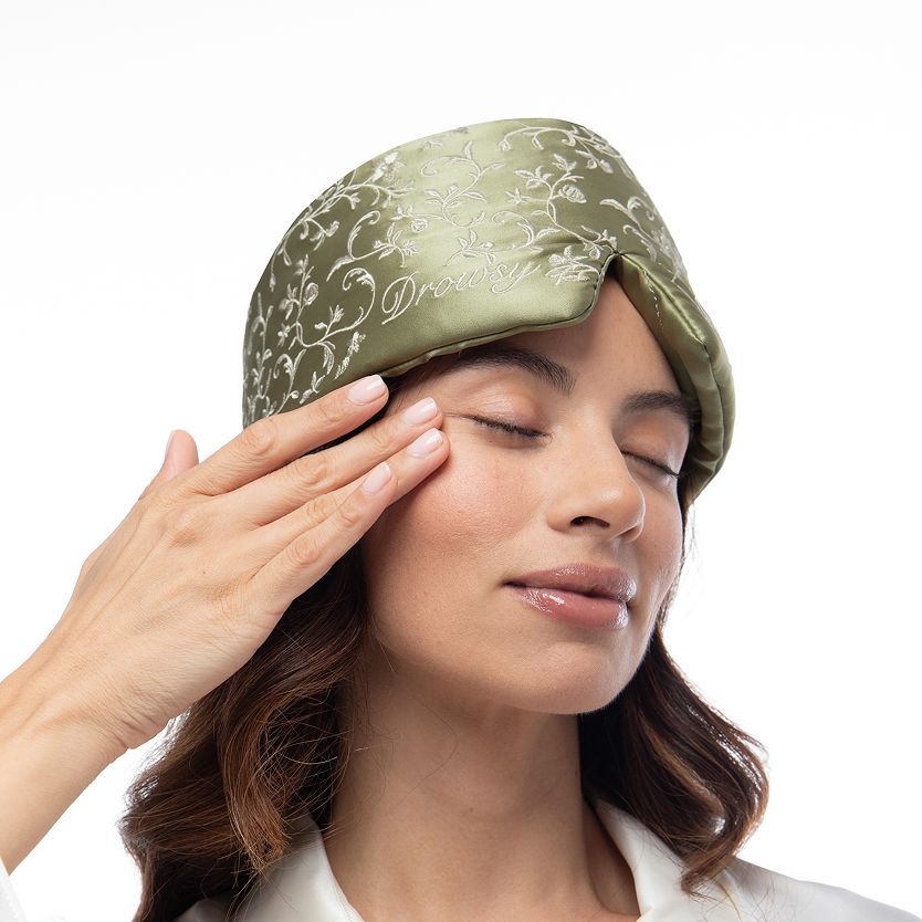 A person wearing a Pride & Prejudice - Sage Sleep Mask, which is sage green with white floral vine embroidery and the word Drowsy.