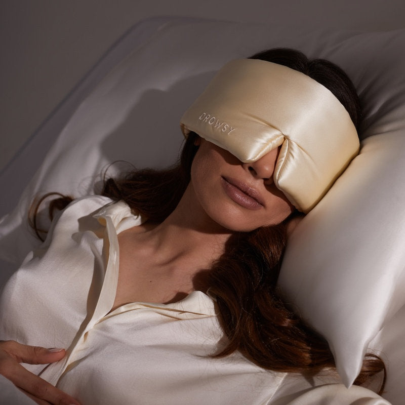 A woman relaxes with her eyes covered, wearing the us.drowsysleepco Champagne Gold silk eye mask and matching pajamas on a satin pillow. The mask features DROWSY embroidery, ensuring ultimate beauty sleep and comfort.