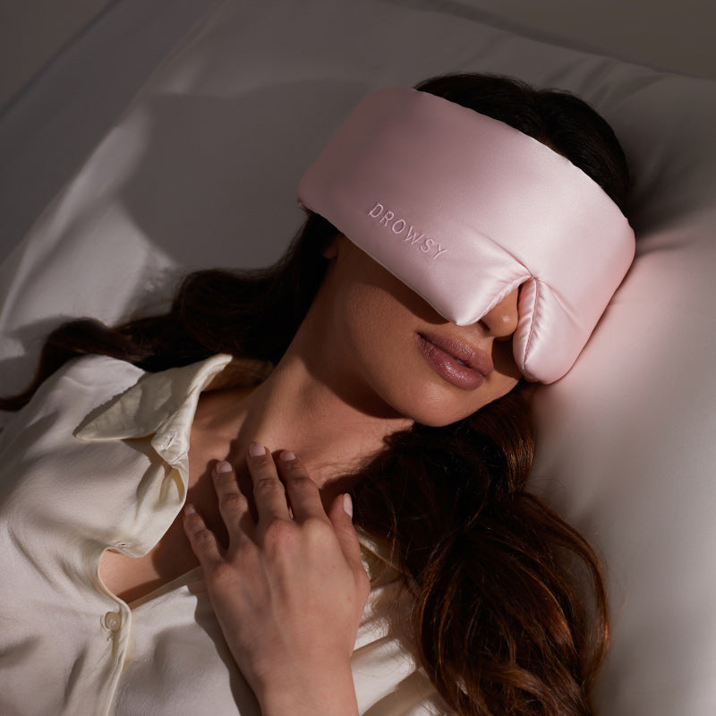 A woman lies comfortably in bed wearing the us.drowsysleepco Petal Pink silk sleep mask, dressed in a cream satin shirt and enjoying the soothing benefits of her blackout eye mask.