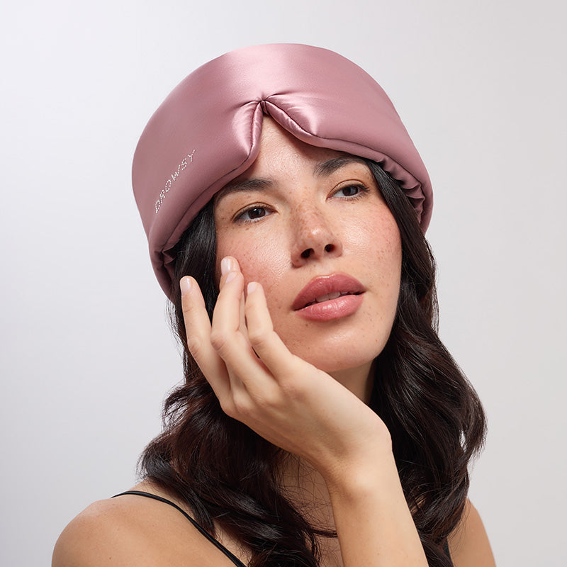 A woman with long dark hair gently touches her face, wearing the Damask Rose head wrap by drowsysleepco with a Swarovski® embellished logo, embodying serene beauty sleep against a neutral background.