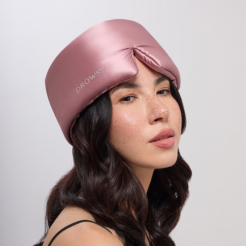 A woman with long dark hair wears the Damask Rose sleep mask cap by drowsysleepco, featuring a Swarovski® embellished logo—shiny pink silk, perfect for beauty sleep—against a plain light background.