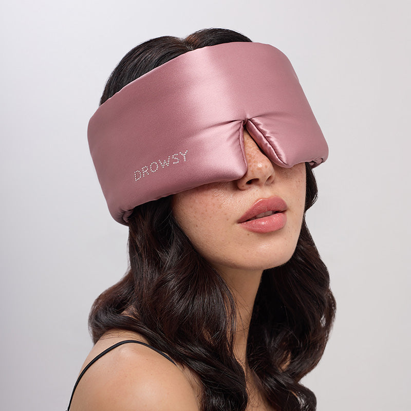 A woman with wavy brown hair wears drowsysleepco’s Damask Rose silk sleep mask, featuring a Swarovski® embellished logo. The wide pink mask covers her eyes and nose as she poses with neutral makeup against a light gray background.