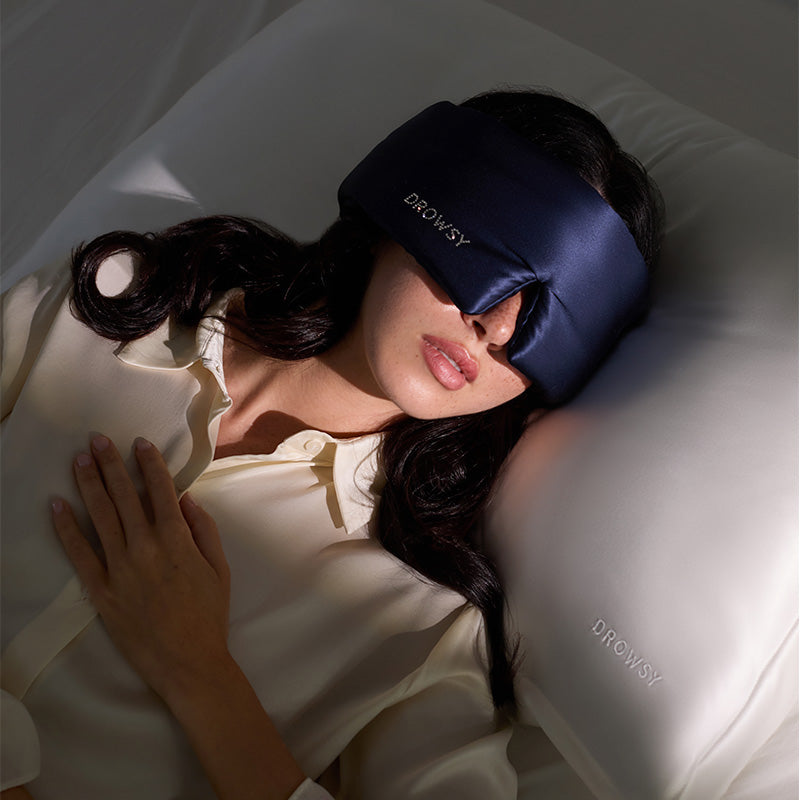A woman lies on her back in bed, wearing a drowsysleepco Midnight Blue sleep mask with a Swarovski® embellished logo. One hand rests on her chest and she uses a white pillow embroidered with "DROWSY.