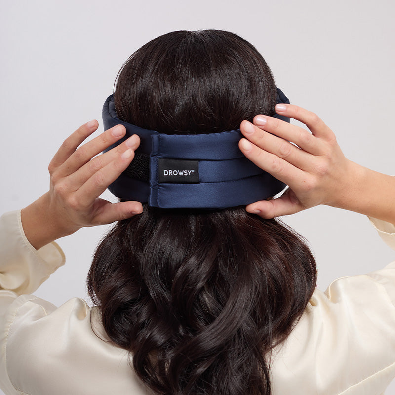 A person with long brown hair adjusts a drowsysleepco Midnight Blue sleep mask with a Swarovski®-embellished logo, wrapping it around the back of their head.