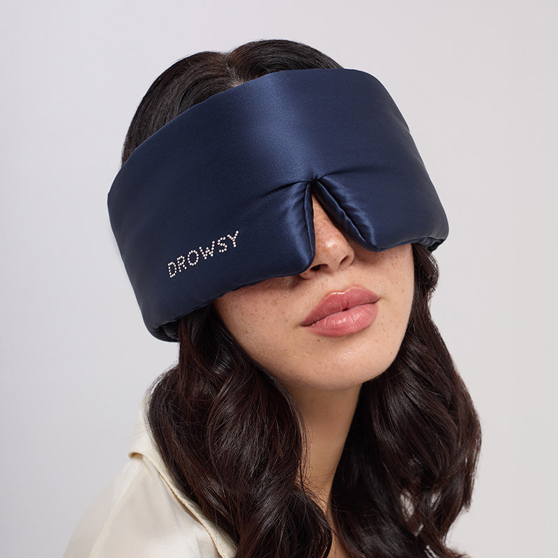 A woman with wavy dark hair wears the drowsysleepco Midnight Blue sleep mask, featuring a Swarovski® embellished logo, covering most of her face. She is dressed in a light-colored top.